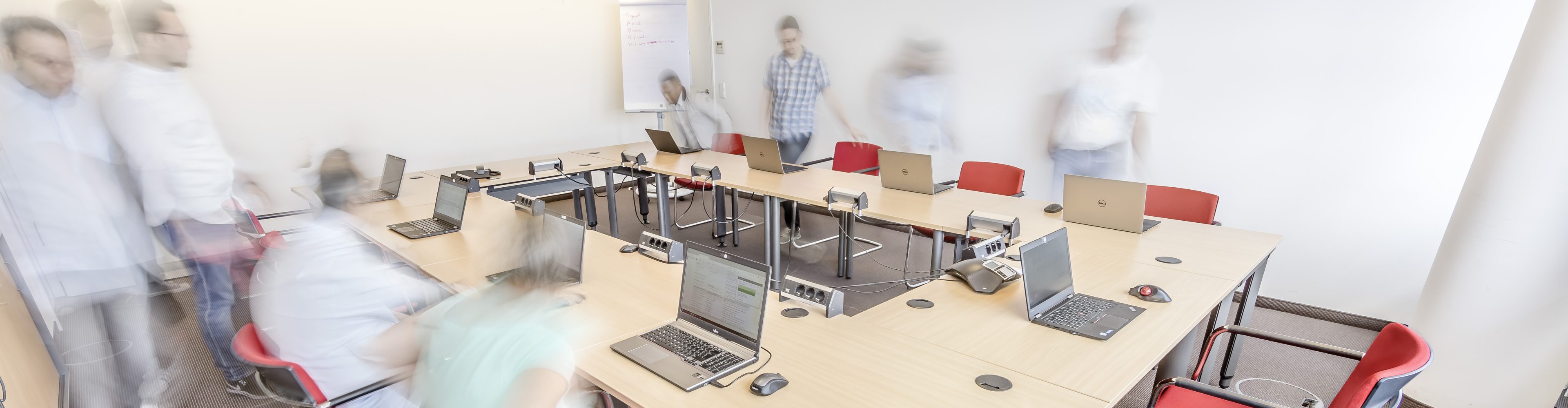 Ein Raum mit quadratisch angeordneten Tischen mit Laptops. Es ist eine Gruppe Personen durchscheinend angedeutet, die sich durch den Raum bewegen.