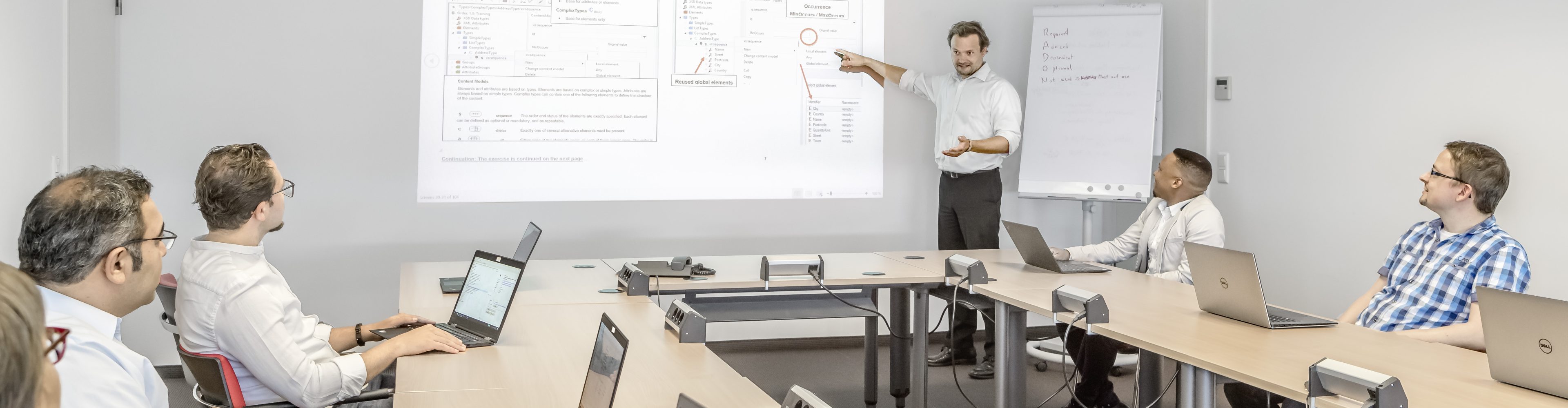 A GEFEG training in which the participants follow the trainer attentively. The trainer explains interrelationships interactively using a flipchart and beamer.