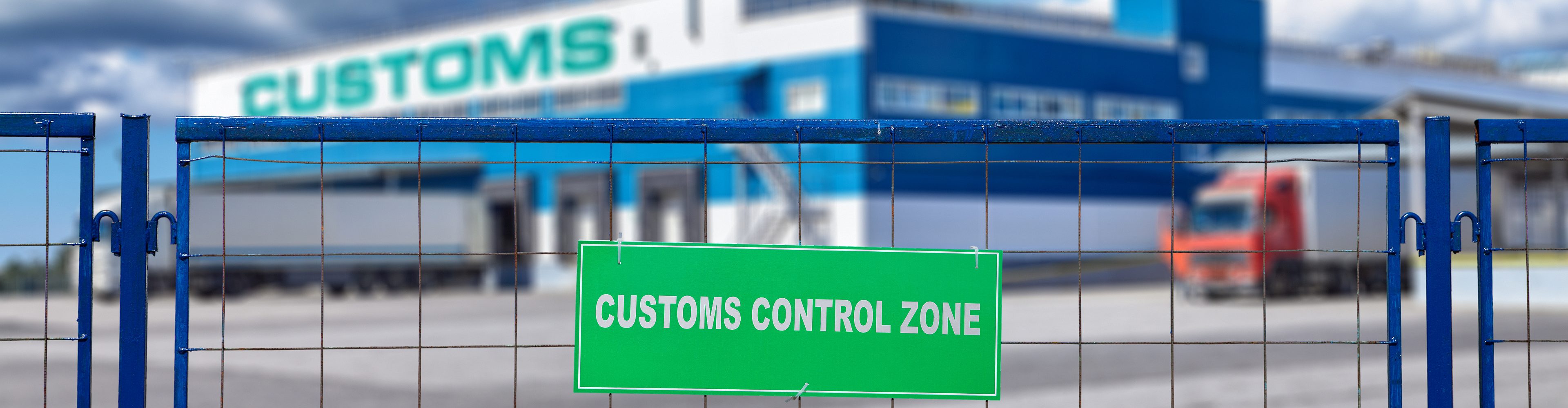 Ein Zaun mit einem Schild, auf dem "Customs Control Zone" steht. Im Hintergrund ist verschwommen ein Abfertigungsgelände zu sehen