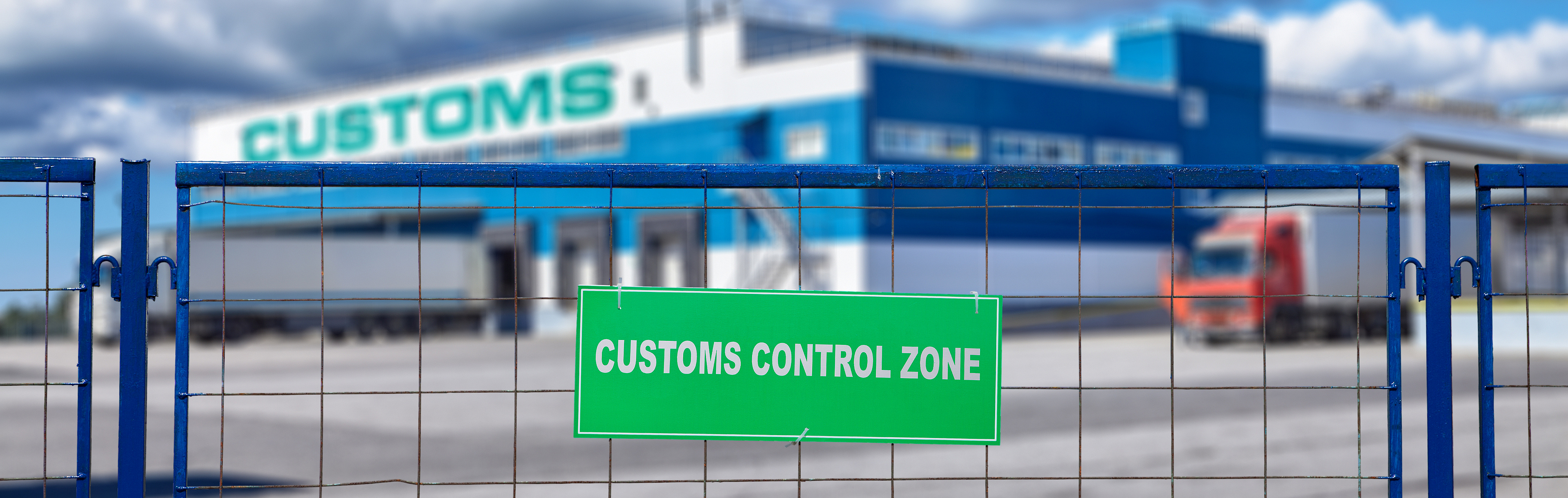 Ein Zaun mit einem Schild, auf dem "Customs Control Zone" steht. Im Hintergrund ist verschwommen ein Abfertigungsgelände zu sehen
